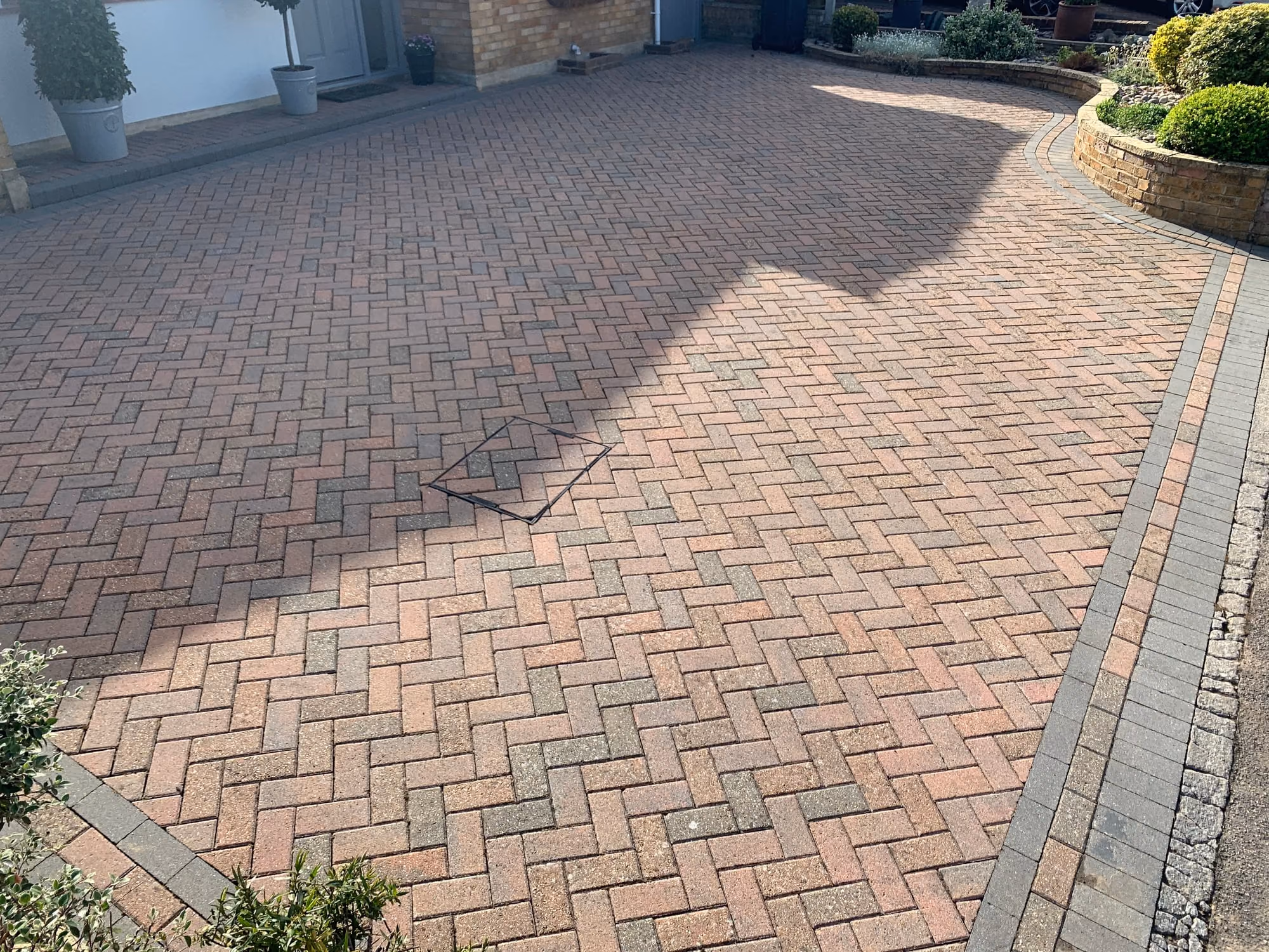 A large herringbone block paving driveway after cleaning, sweeping round a raised planted bed in front of a white rendered house