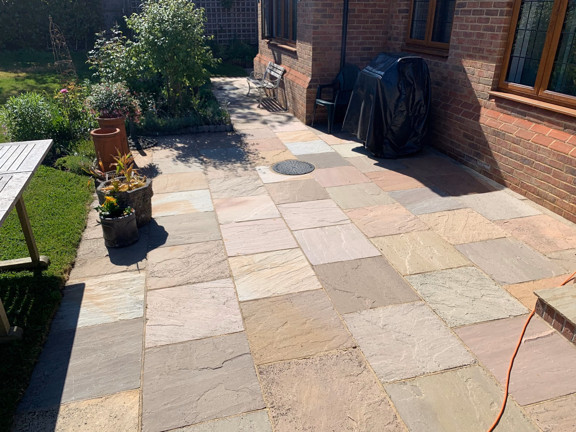 Cleaned Indian sandstone patio running along the back of a red brick house in full sun, with planters and a garden bench at the edge